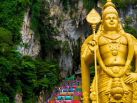 A golden statue of a divine figure stands proudly in front of a majestic Batu Caves mountain backdrop.